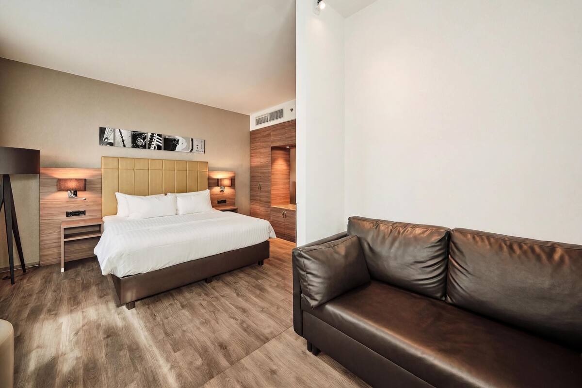 Una habitación de hotel moderna con una cama grande, un sofá de cuero, una obra de arte en la pared y una lámpara de pie.