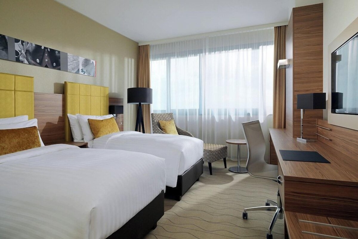 A hotel room with two beds, a desk, a chair, a lamp, and a large window with curtains.