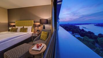 Relax in a stylish room with a large bed, bedside table, and a comfortable armchair by the window overlooking a serene river view.
