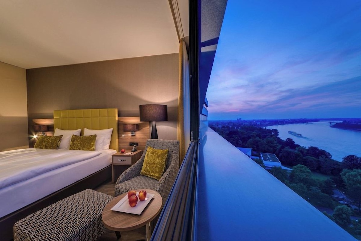 A hotel room with a bed, a sofa, a small table with apples, and a view of a river at dusk.