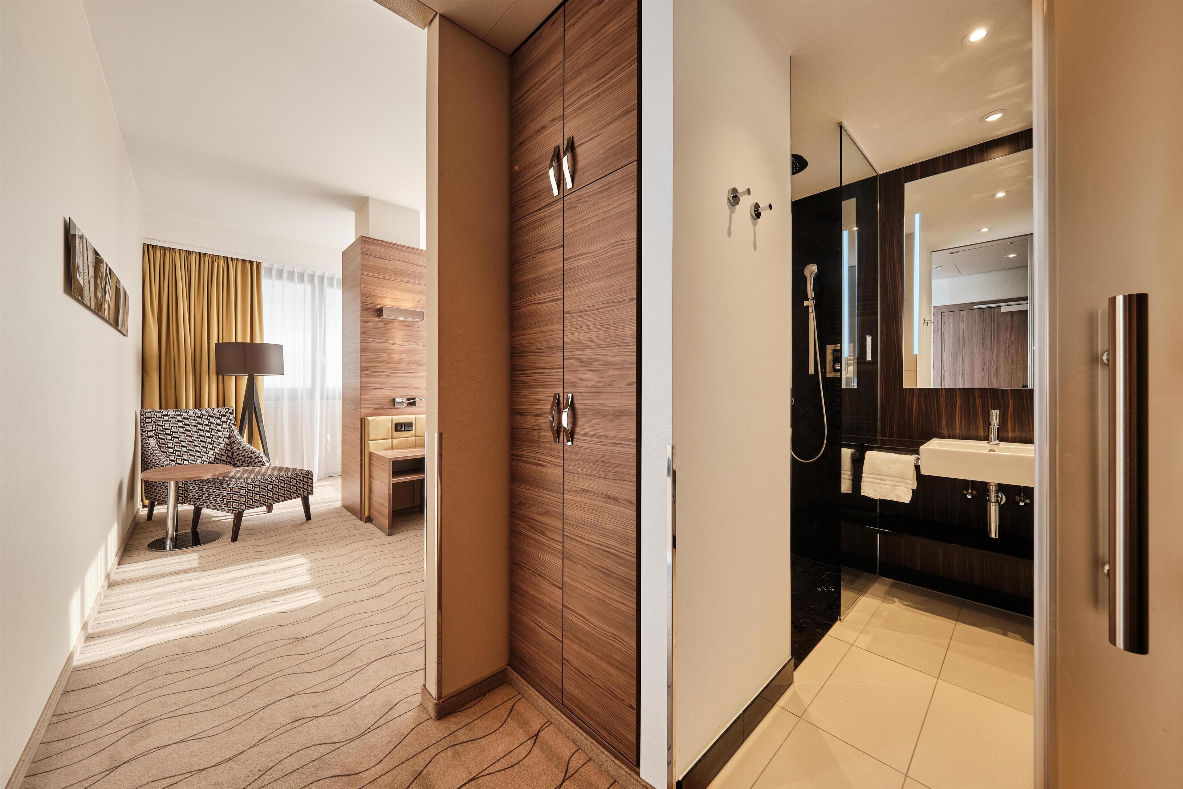 A hotel room with a bed, a desk, a chair, a wardrobe, a bathroom with a sink and mirror, and a shower.