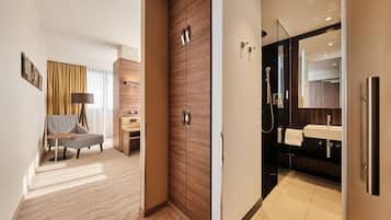 A hotel room with a bed, a desk, a chair, a wardrobe, a bathroom with a sink and mirror, and a shower.