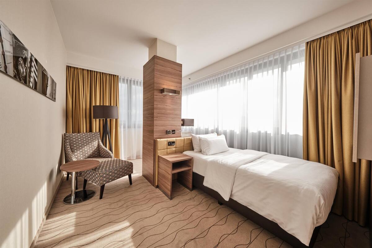 Habitación de hotel con cama, escritorio, silla, lámpara y un amplio ventanal con cortinas.