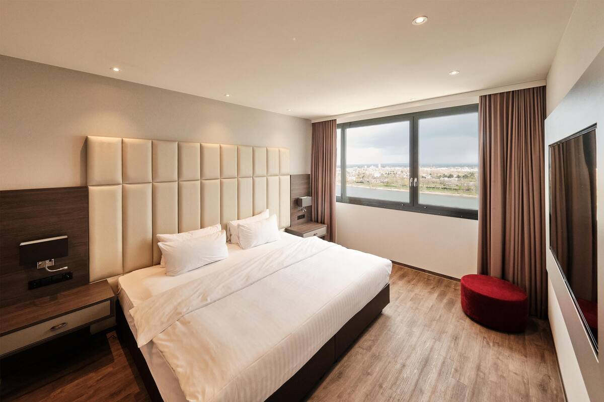 Una habitación de hotel moderna con una cama grande, una mesita de noche, un televisor y un ventanal con vista a la ciudad.