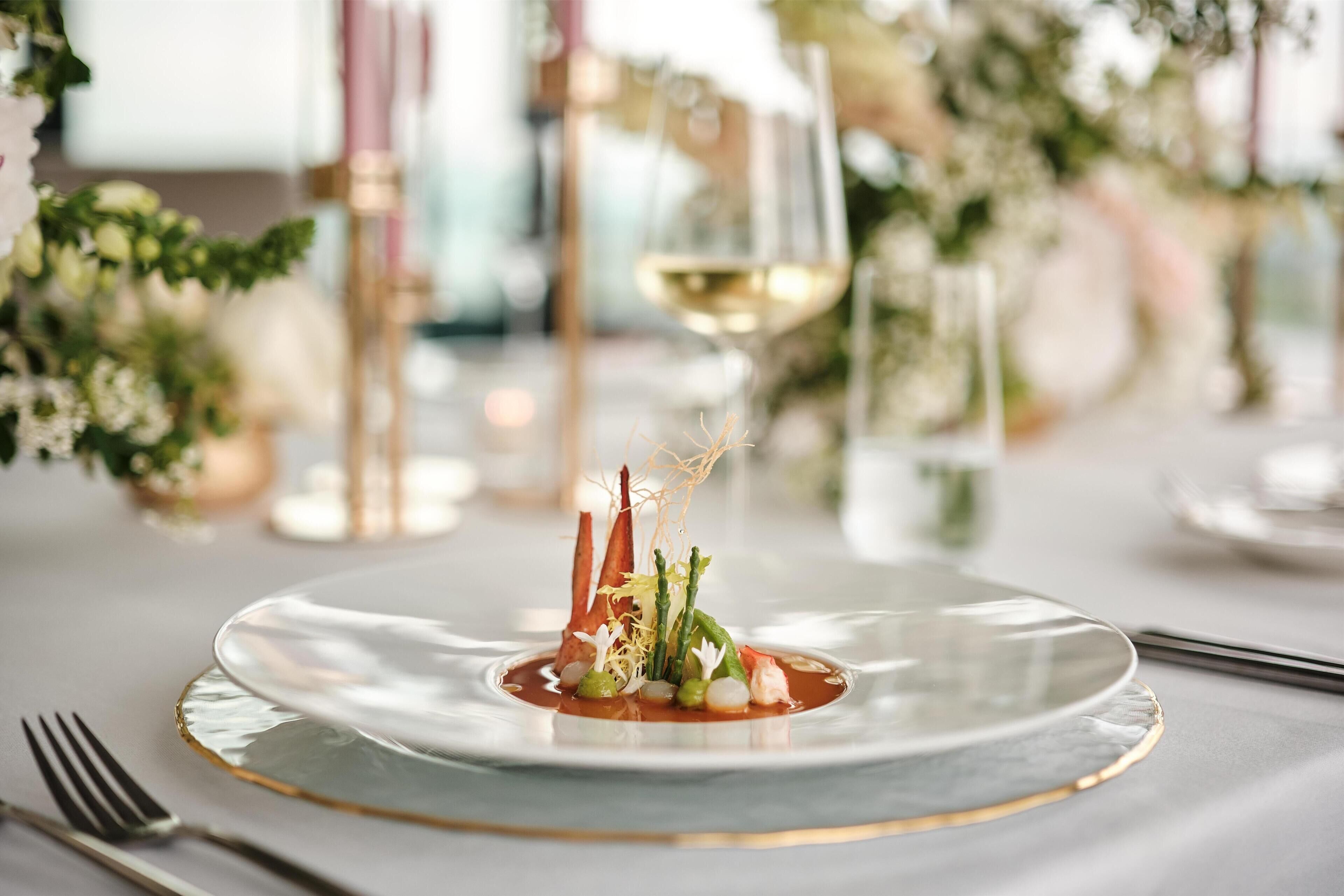 A dining table with a glass plate, a fork, and a glass of white wine.