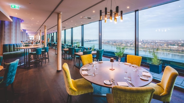 Un restaurante moderno con vistas panorámicas a un río y al paisaje urbano, que cuenta con mesas redondas, asientos cómodos y un techo alto con iluminación empotrada.