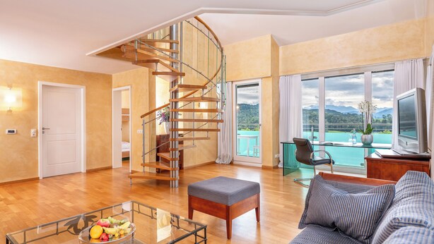 A spacious living room with a wooden spiral staircase, a large window with a view of mountains, a sofa, and a glass coffee table with fruit. - Allgäu Stern Hotel (Sonthofen)