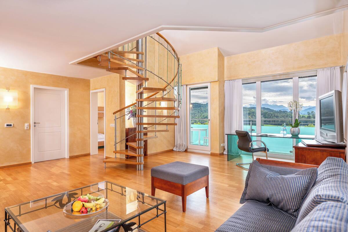 Ein geräumiges Wohnzimmer mit einer Holzspirale, einem großen Fenster mit Bergblick, einer Couch und einem Glastisch mit Obst.