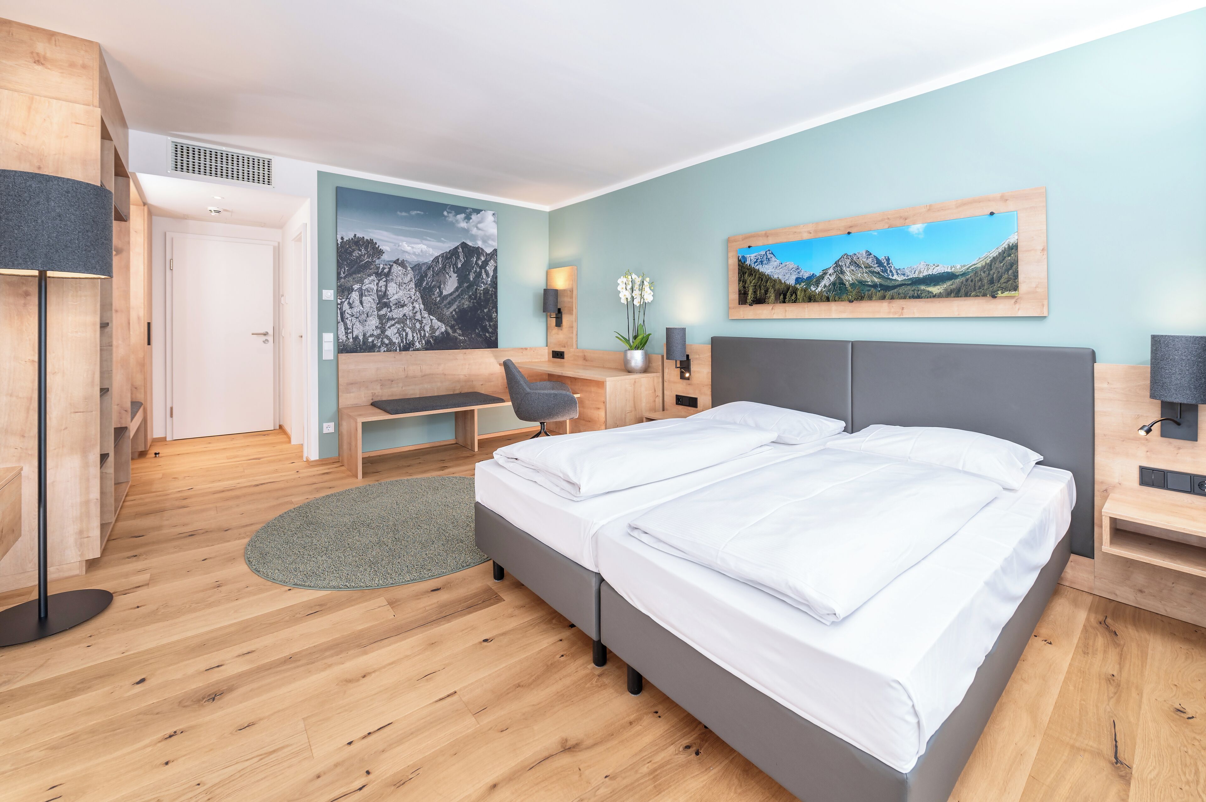 A modern hotel room with a wooden floor, a double bed, a desk, a chair, and two paintings on the walls.