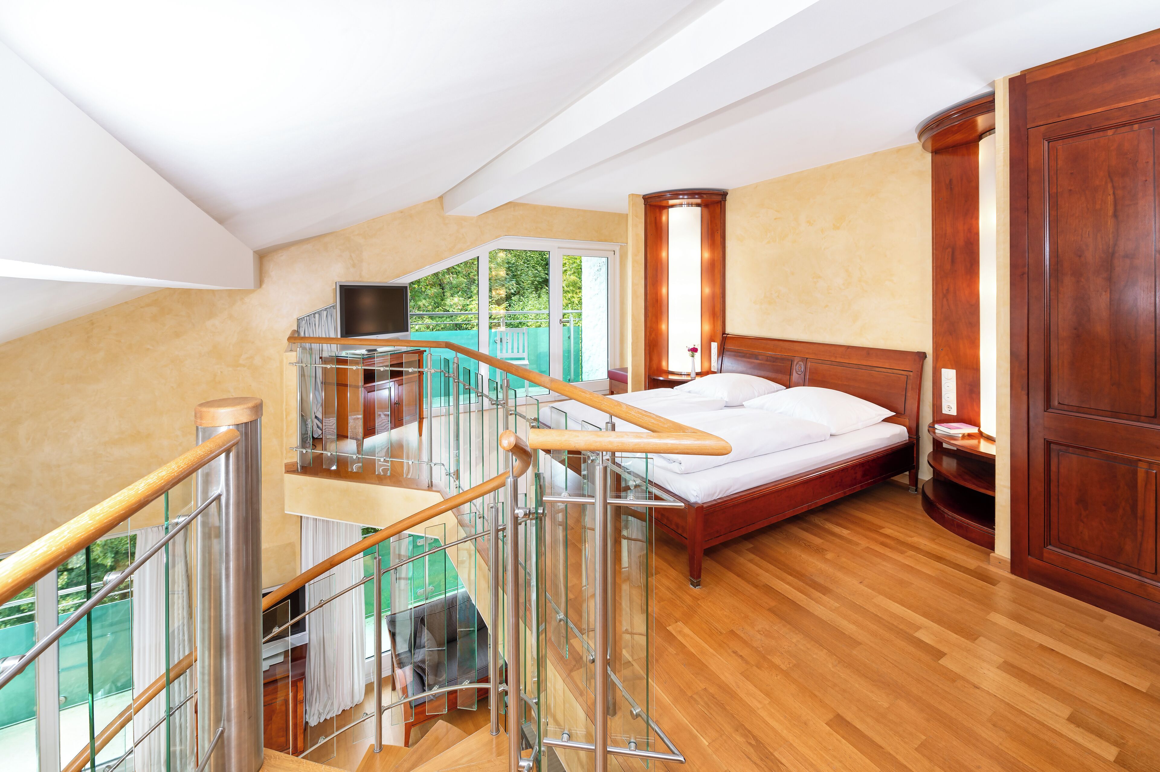 A bedroom with a wooden bed, a glass staircase, and a view of greenery outside.
