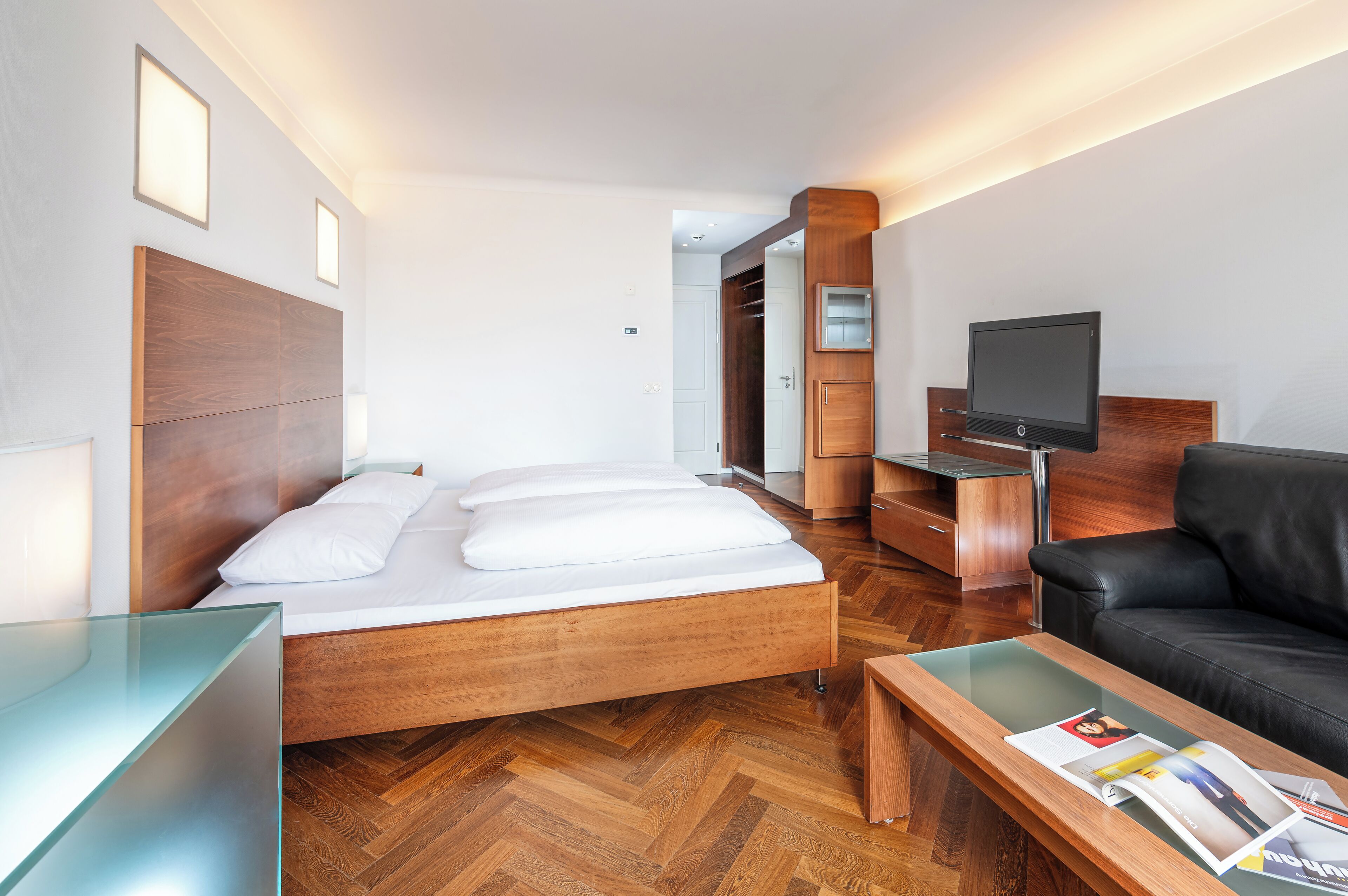 Ein modernes Hotelzimmer mit einem hölzernen Bett, einer schwarzen Ledersofa, einem Fernseher und einem Glastisch.