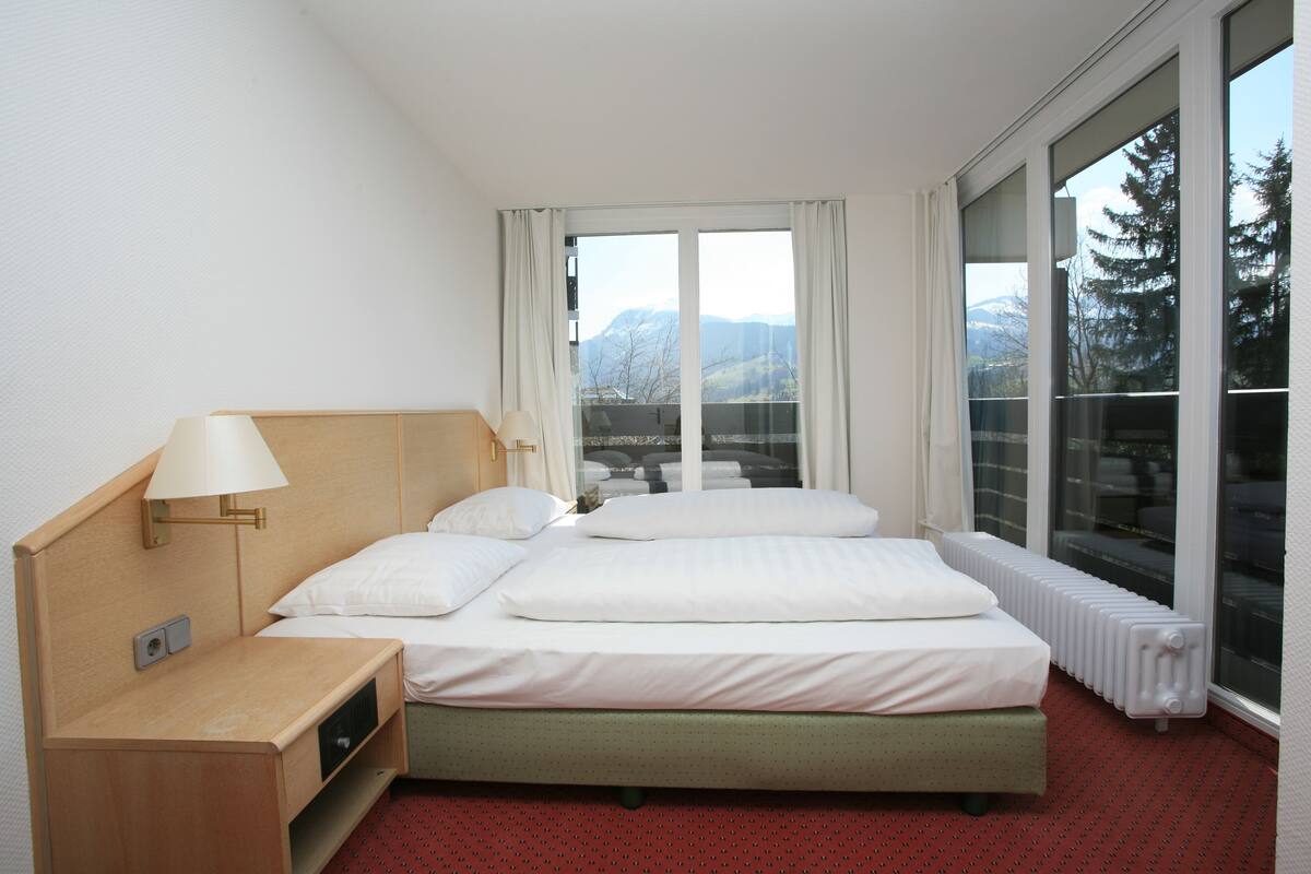 Ein Hotelzimmer mit Bett, Nachttisch, Lampe, einem Fenster mit Bergblick und einer Heizung.