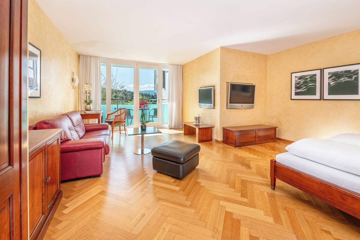 Ein Hotelzimmer mit Holzdielen im Fischgrätmuster, einem roten Ledersofa, einem hölzernen Bett, einem kleinen Tisch mit Pflanze und einer Glastür zum Balkon.