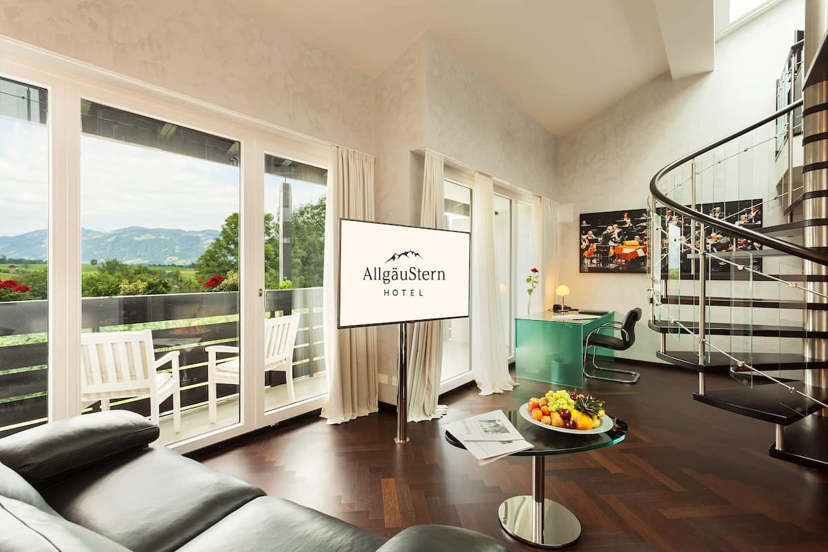 Ein Hotelzimmer mit großem Fenster, einer Couch, einem Esstisch mit Obst und einem Schild mit der Aufschrift "AllgäuStern Hotel".