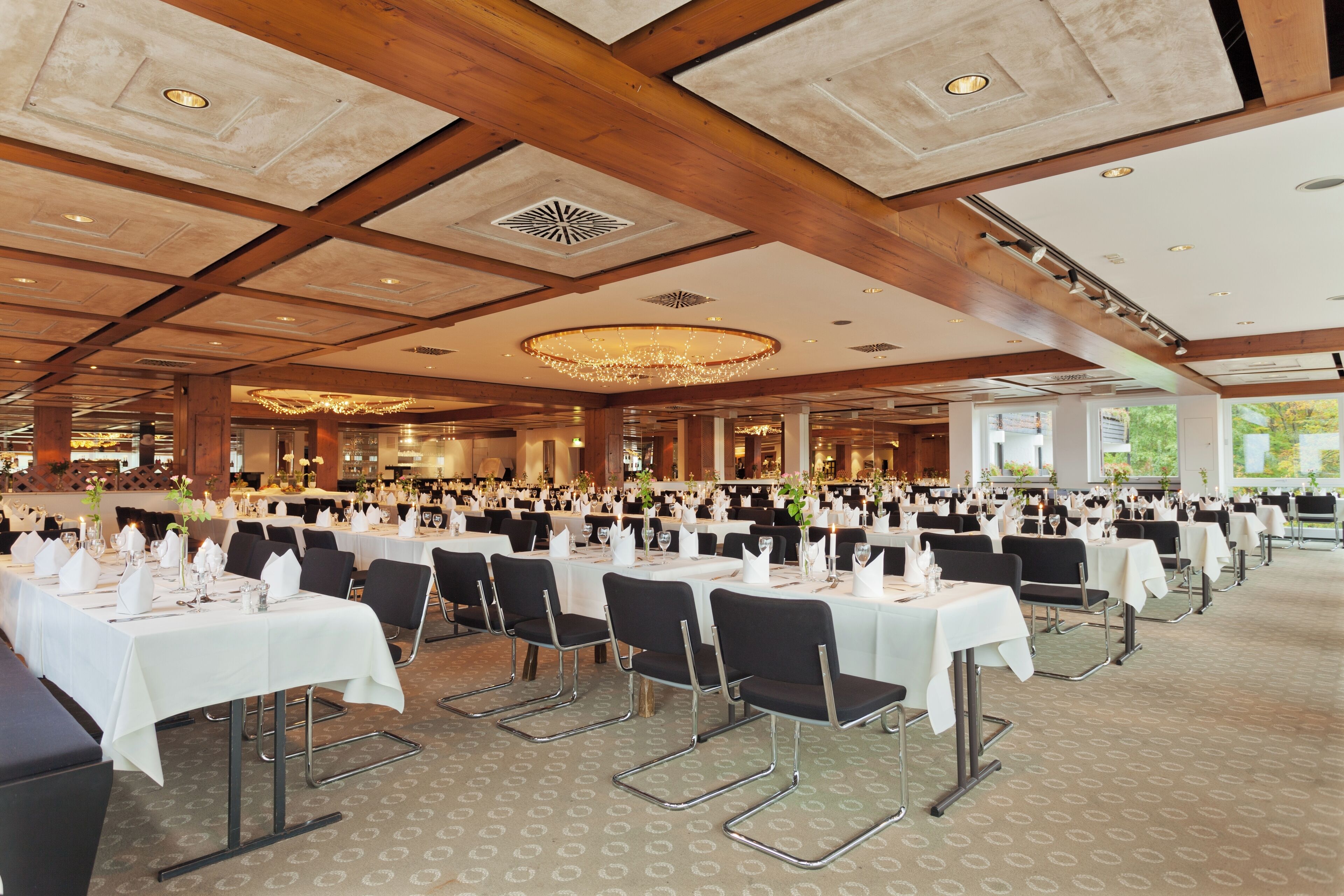A large banquet hall with multiple tables set for dining, featuring a high ceiling with wooden beams and recessed lighting.
