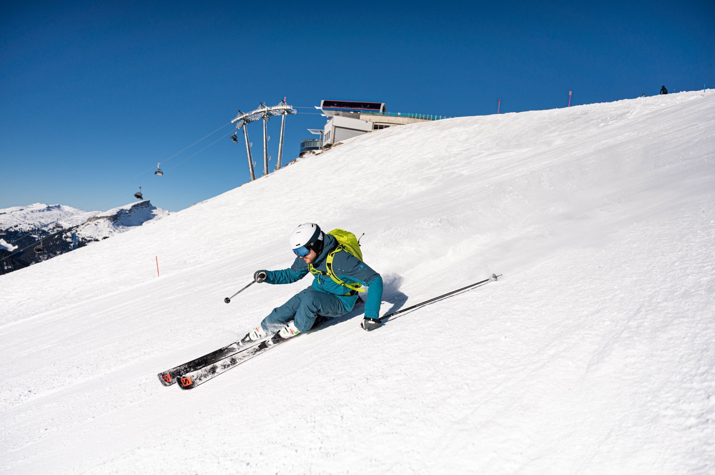 Eine Person fährt auf Skiern einen verschneiten Hang hinunter, im Hintergrund sind Skilifte zu sehen.