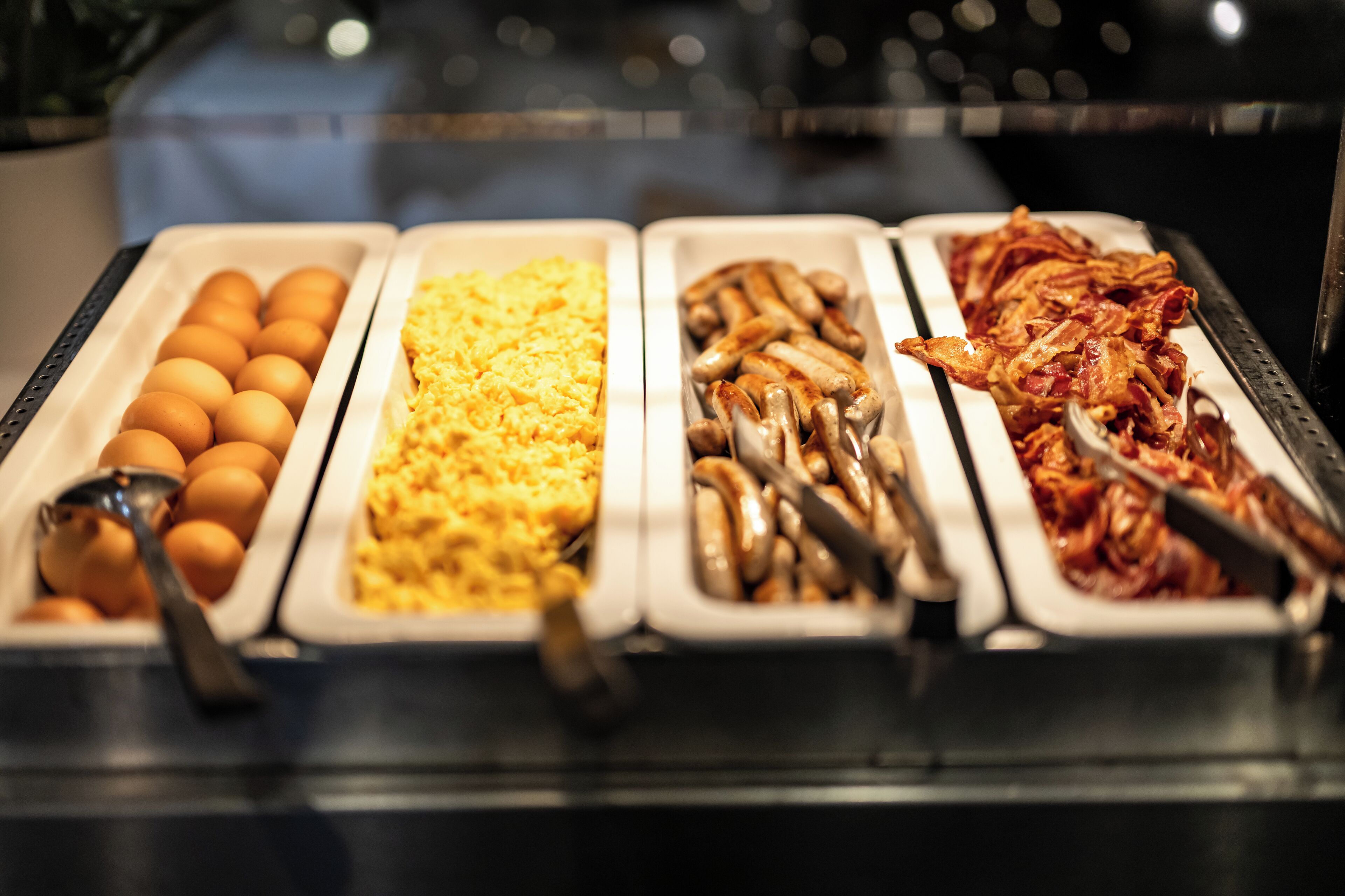 A buffet with trays of eggs, scrambled eggs, sausages, and bacon.