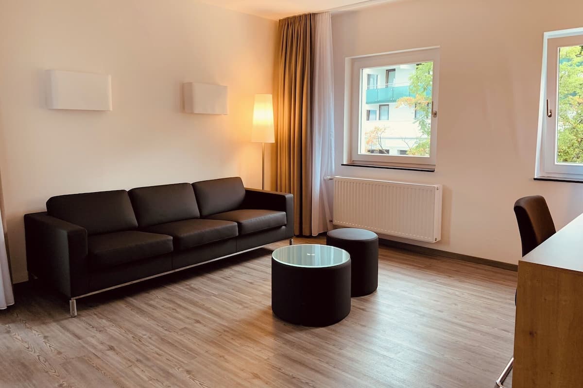Ein modernes Zimmer mit einer schwarzen Couch, einem kleinen runden Tisch und einem Fenster mit Blick auf Gebäude und Bäume.