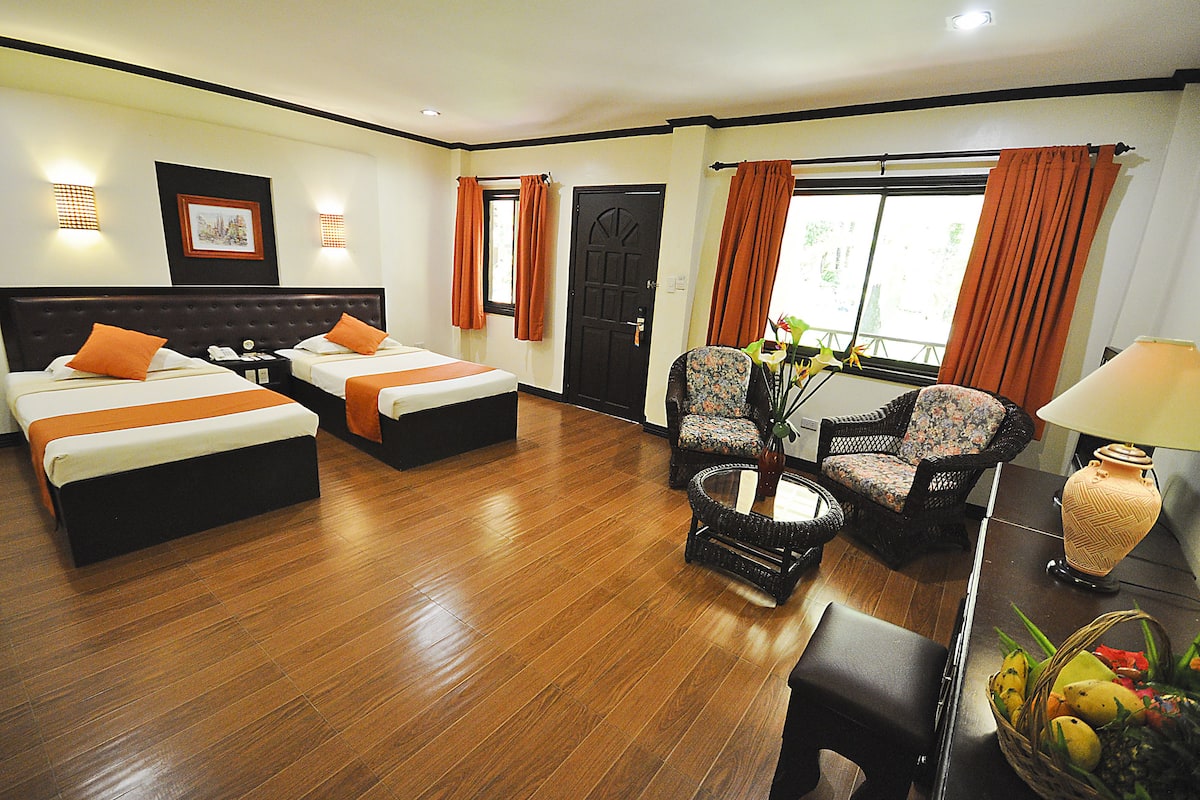 A hotel room with two beds, a sitting area with a chair and table, a lamp, and a window with curtains.