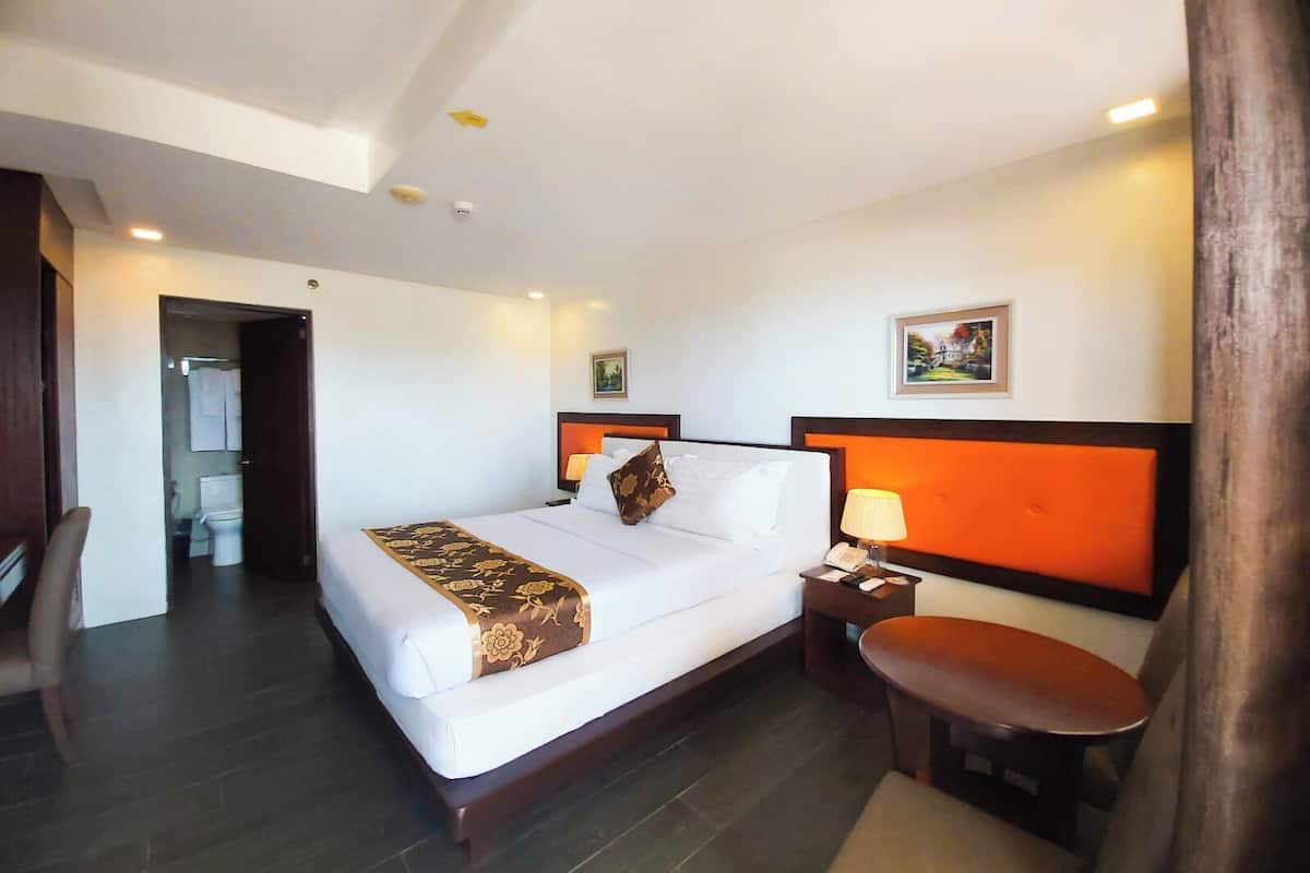 A hotel room with a bed, bedside table, chair, and a painting on the wall.