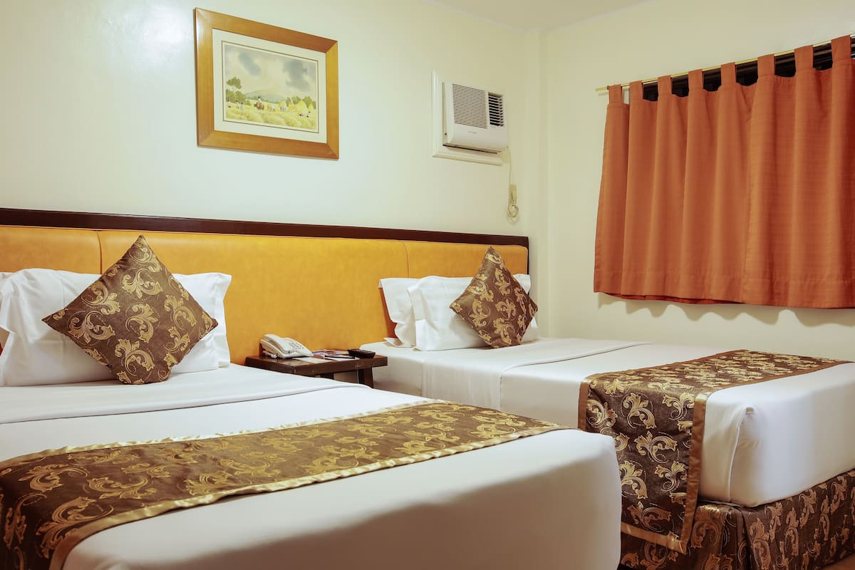 A hotel room with two beds, each with a patterned bedspread, white bedding, and decorative pillows. There is a nightstand with a telephone and an air conditioning unit mounted on the wall.