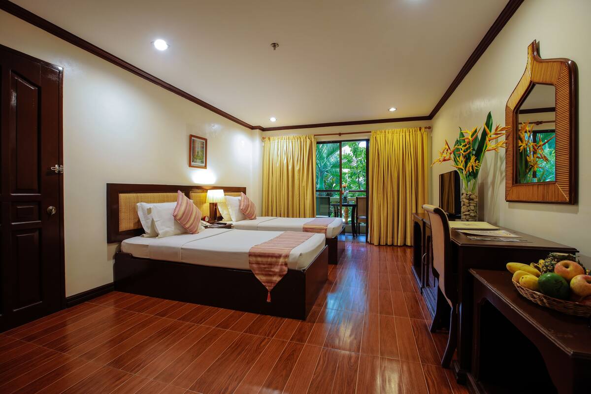 A hotel room with two beds, a desk with a fruit basket, a mirror, and a view of greenery outside.