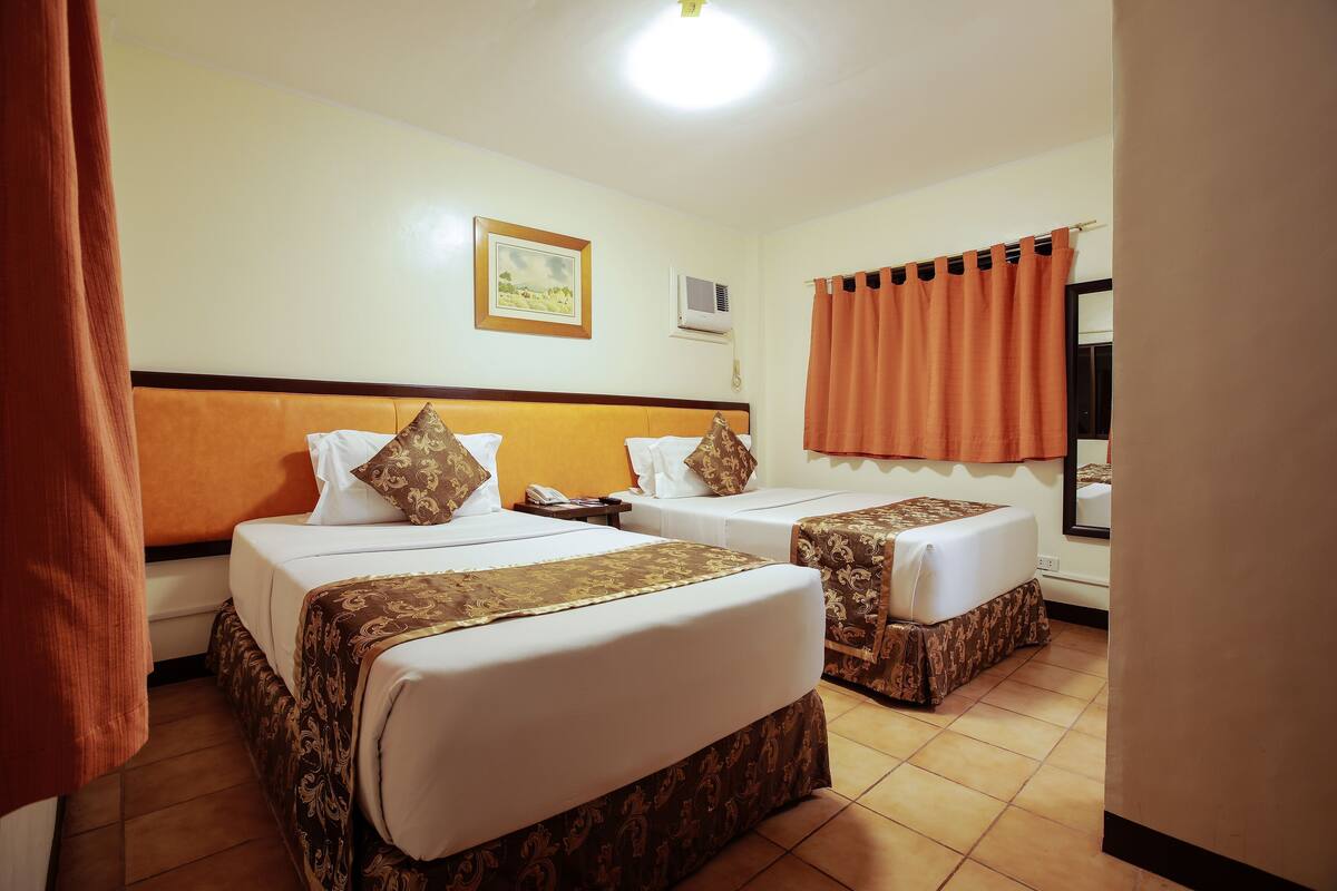 A hotel room with two beds, a small table, a wall-mounted phone, and a window with curtains.