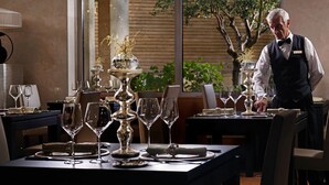 A well-set dining table with multiple wine glasses and a centerpiece, a waiter in uniform, and a view of greenery outside. - Divani Escape- Adults only (Vari-Voula-Vouliagmeni)