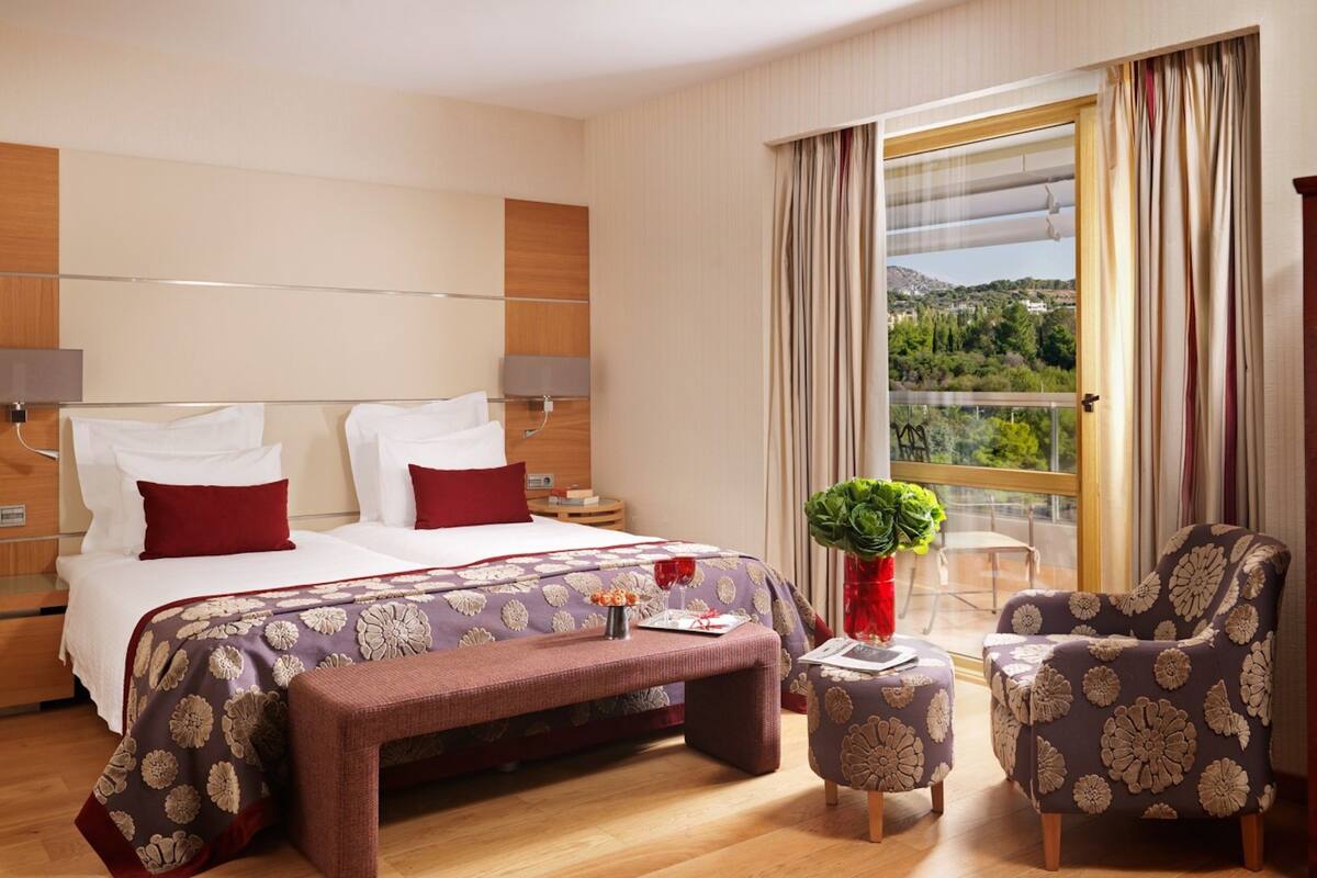 A hotel room with a large bed, a bench, two armchairs, a small table, a plant, and a view of a mountainous landscape.
