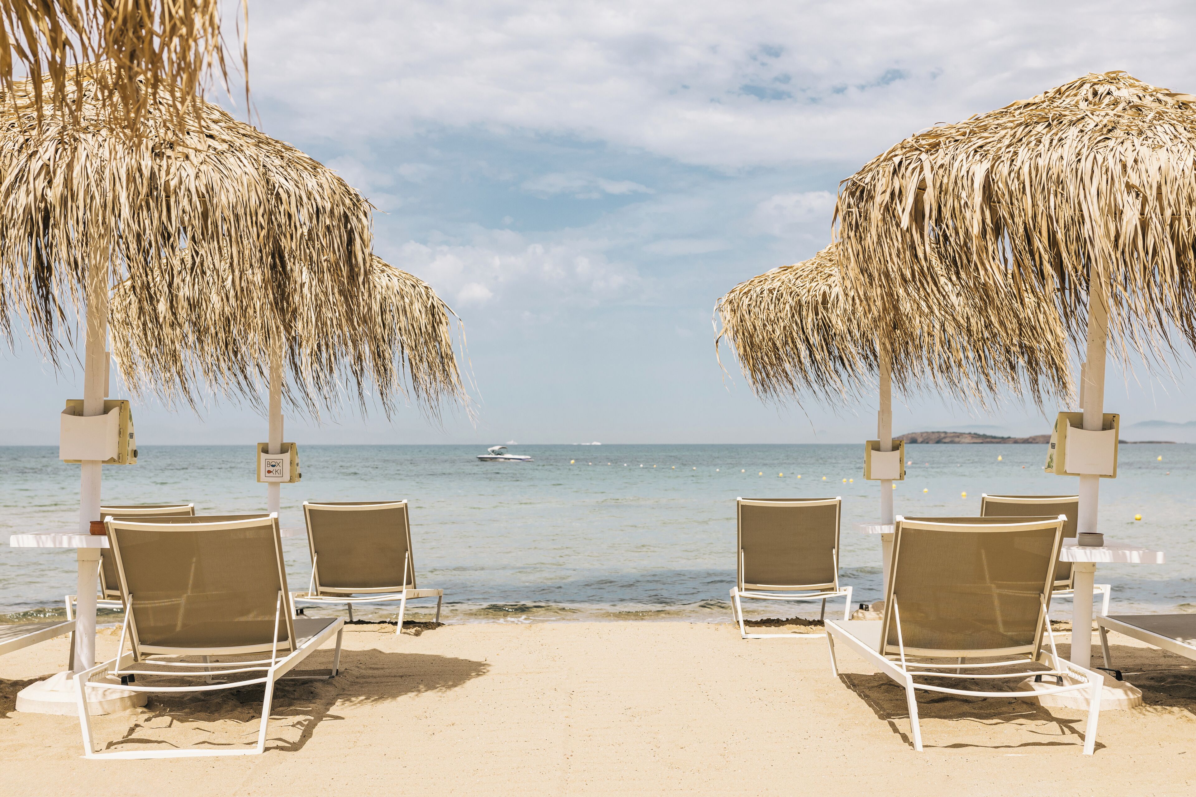 A beach with four white lounge chairs under straw umbrellas, facing the sea.