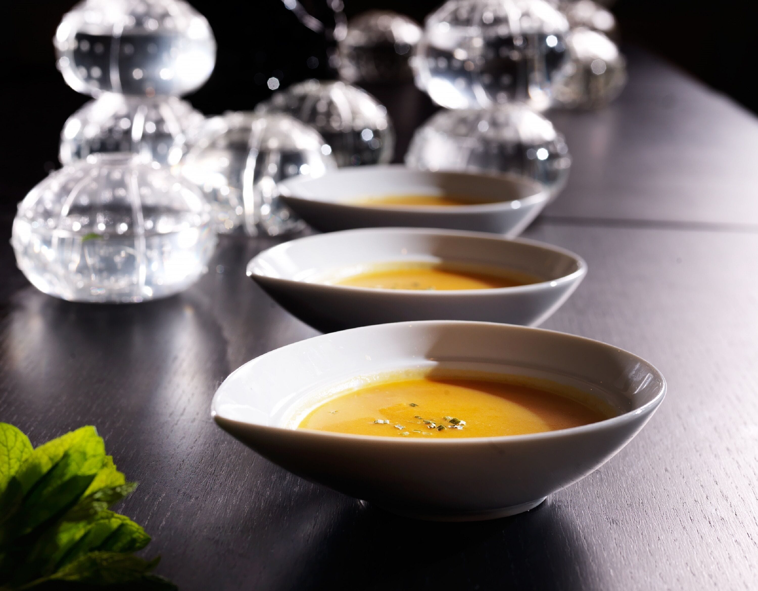Tres tazones blancos de sopa sobre una superficie oscura, con botellas de vidrio decorativas al fondo.