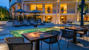 An outdoor pool area with lounge chairs and tables, surrounded by a modern building with large windows. - Divani Escape- Adults only (Vari-Voula-Vouliagmeni)