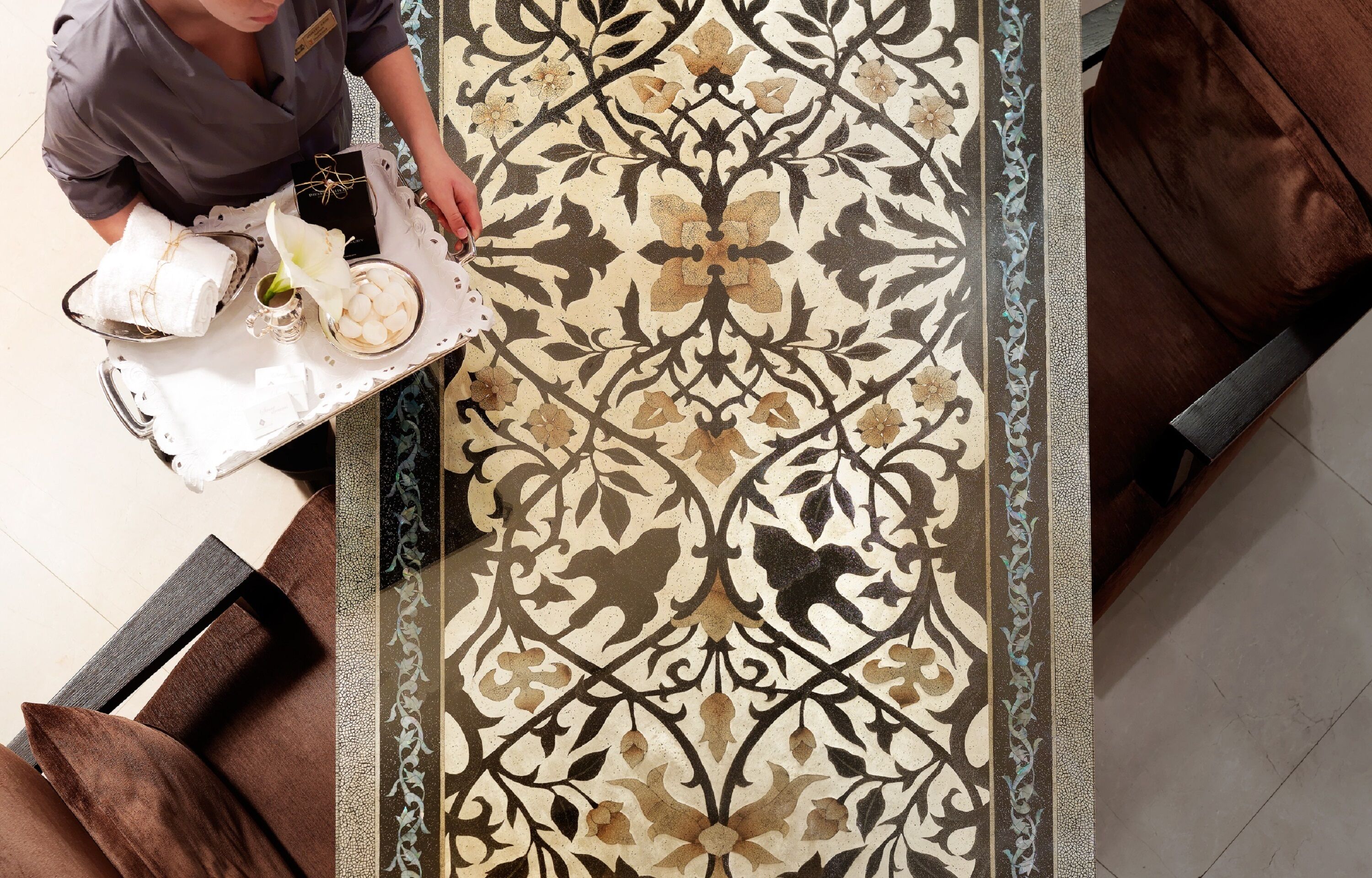 Une personne dispose des serviettes et une fleur sur un plateau dans une pièce dont le sol est recouvert d’un tapis à motifs et où se trouvent des meubles marron.
