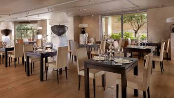 A spacious dining area with multiple tables set for dining, elegant lighting fixtures, and large windows offering views of greenery.
