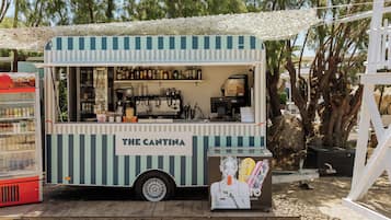 Un camión de comida llamado 'The Cantina' con un diseño a rayas, ubicado en una cubierta de madera con arena de fondo y árboles a lo lejos.