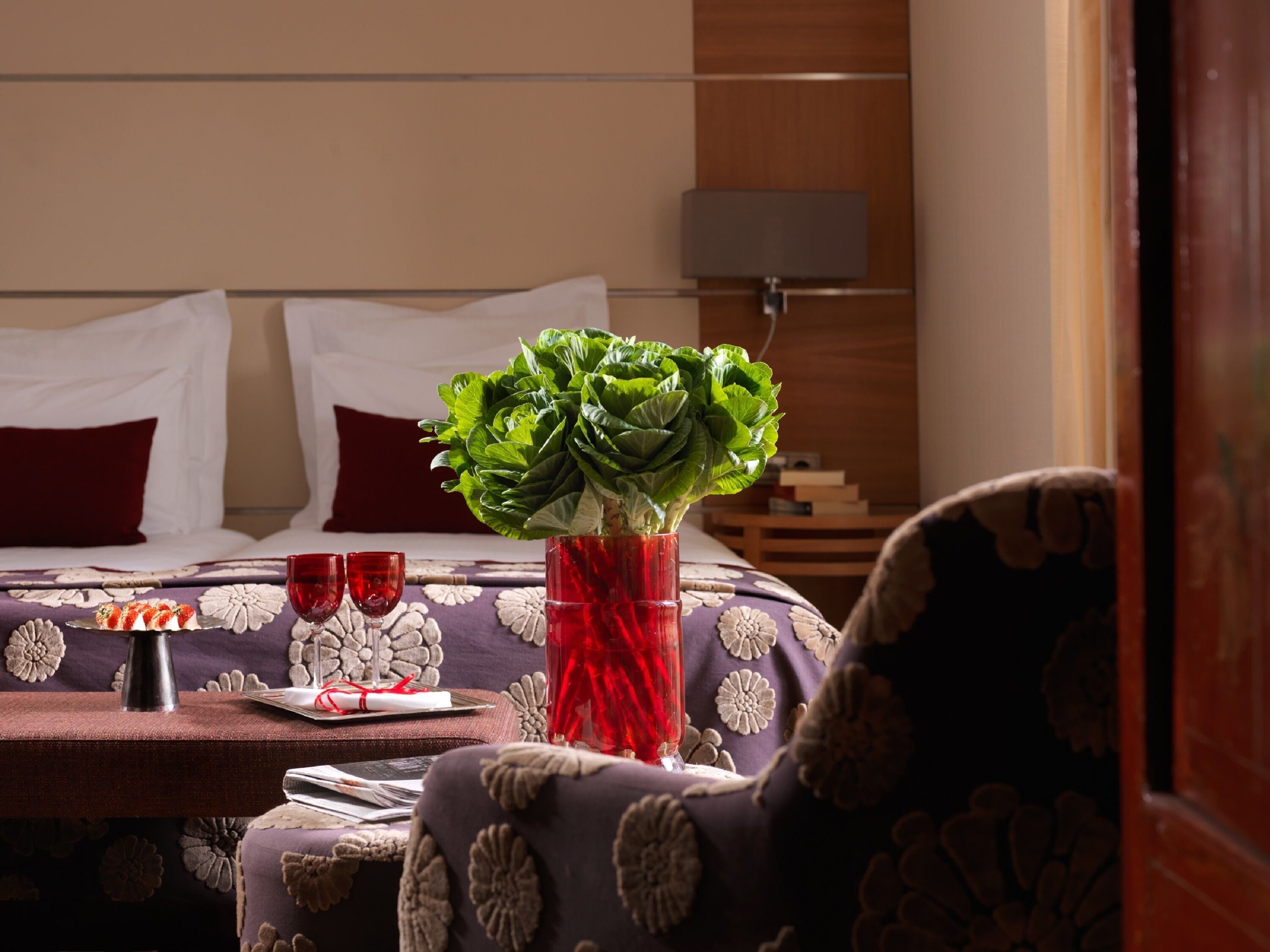 A neatly arranged bedroom with a bed, a bedside table with a vase of flowers, two glasses, and a plate with a dessert.