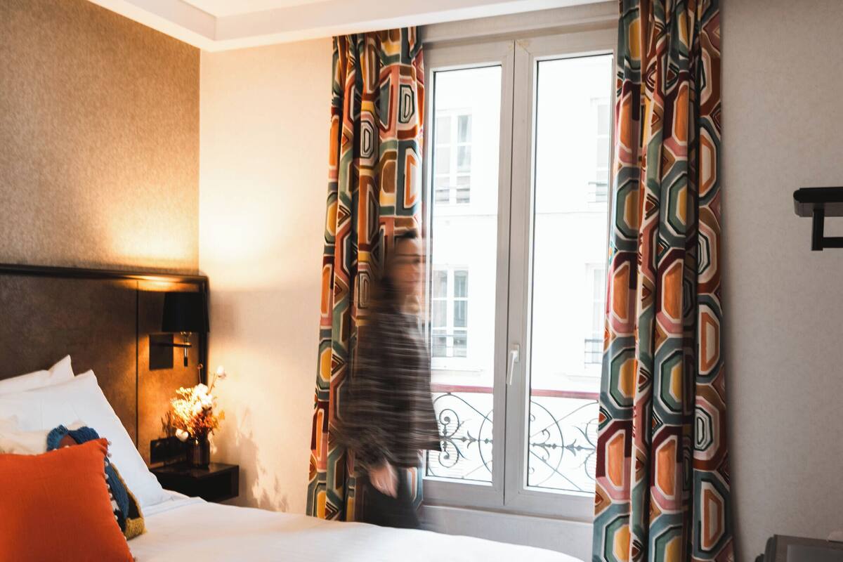 A hotel room with a large bed, a nightstand with a lamp, a window with curtains, and a view of buildings outside.