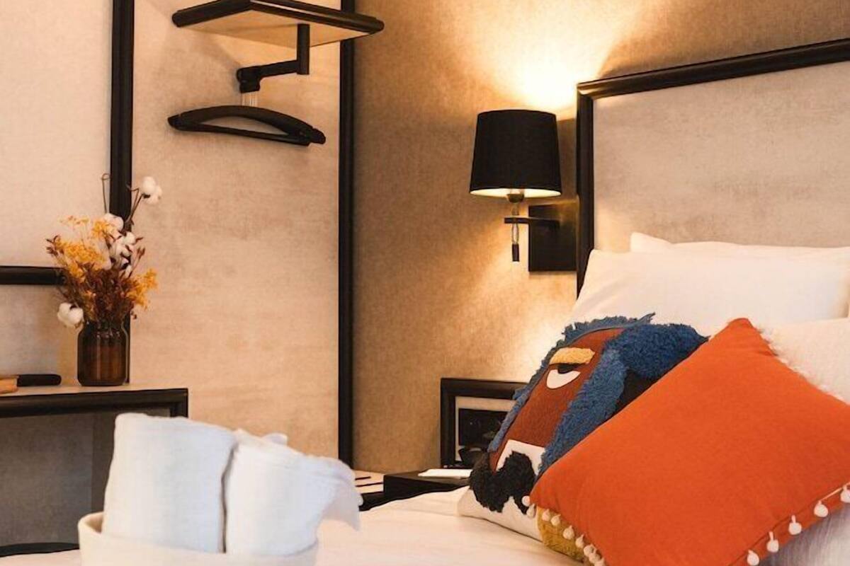 A modern hotel room with a bed, a bedside table with a lamp, a wall-mounted shelf, and a vase with flowers.
