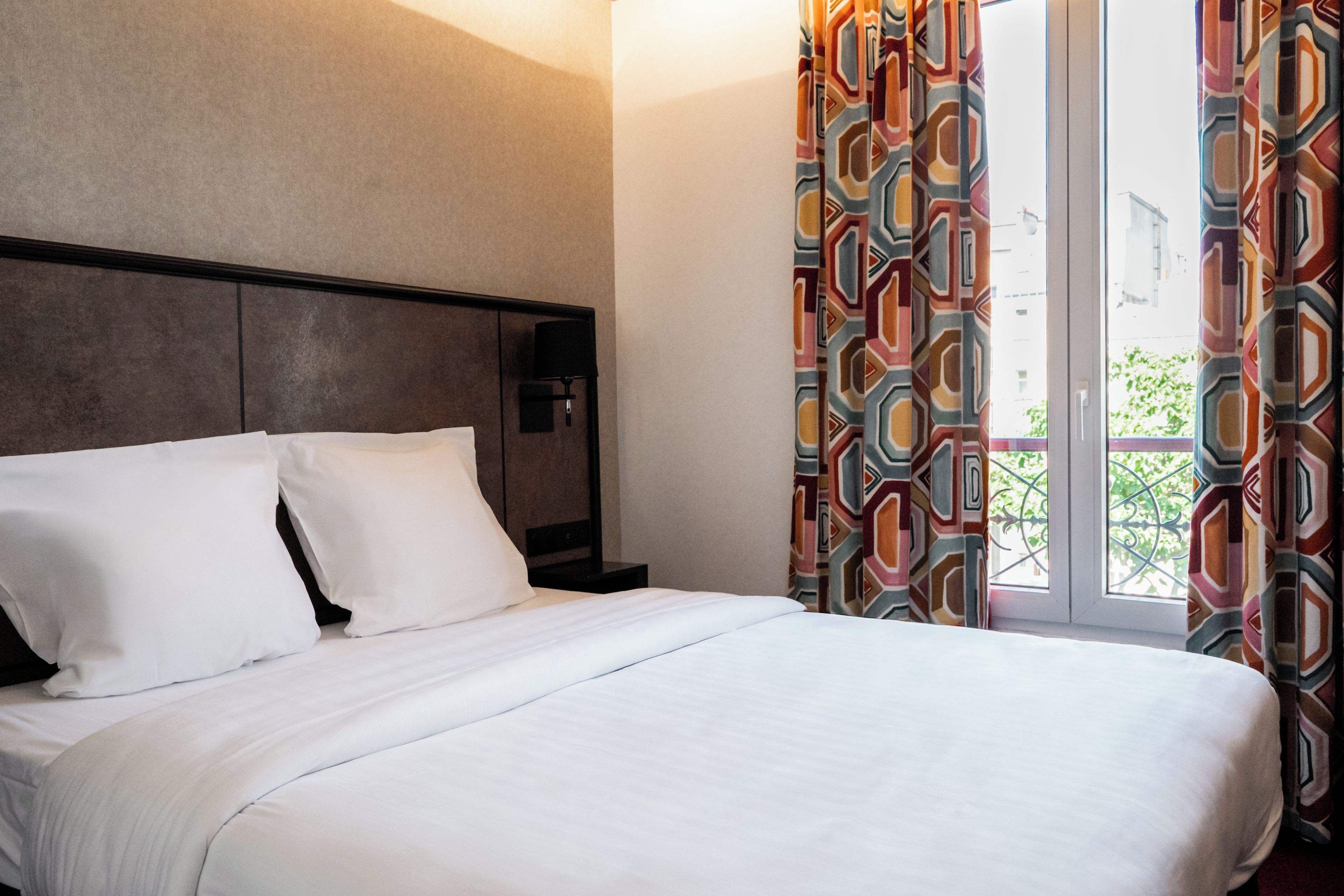 Quarto de hotel com cama, mesa de cabeceira, abajur e janela com cortinas.
