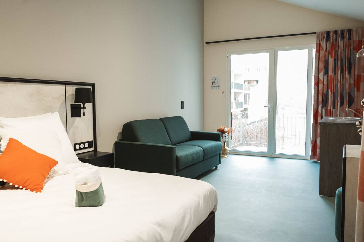 A hotel room with a bed, a green sofa, a small table with a vase, and a view through a glass door.