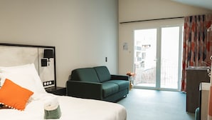 Habitación de hotel con una cama, un sofá verde, una mesita con un florero y vistas a través de un ventanal.