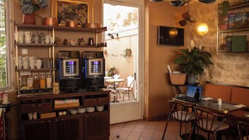 Interior de café com máquina de café, prateleiras com xícaras e potes, mesa com cadeiras e televisão na parede.