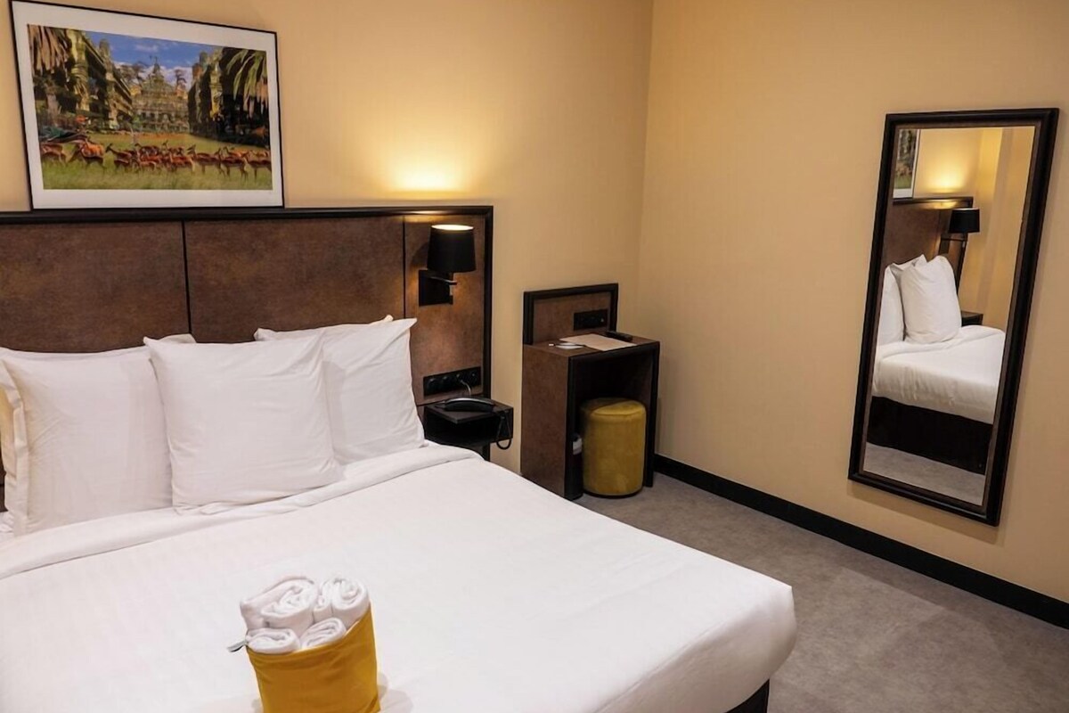 A hotel room with a bed, a bedside table, a lamp, a mirror, and a painting on the wall.