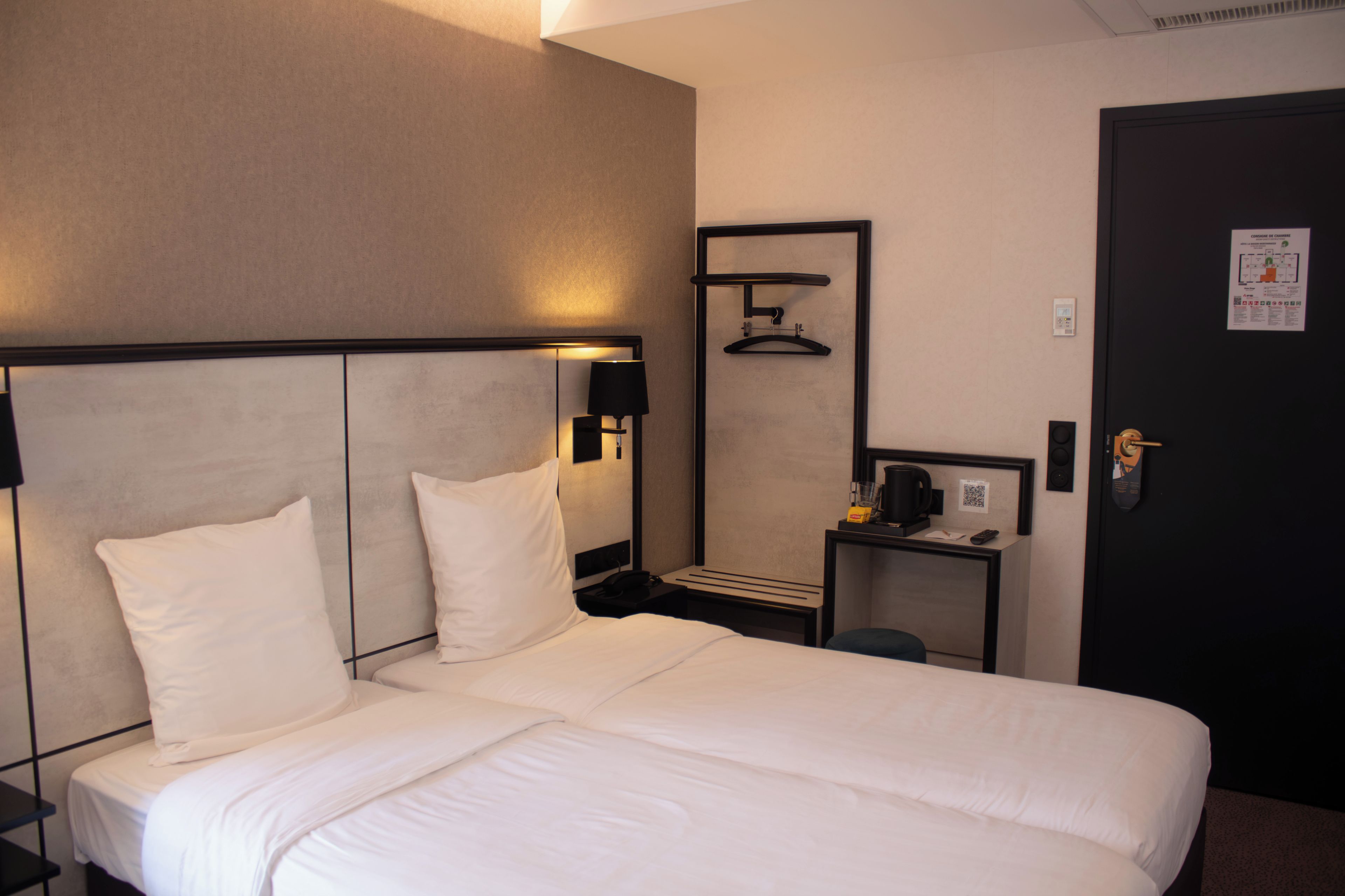 Ein Hotelzimmer mit einem großen Bett, einem Nachttisch mit Lampe, einem kleinen Tisch mit Kaffeemaschine und einer Tür mit Zimmerschlüssel und QR-Code.