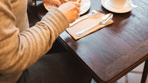 Una persona sentada en una mesa de madera con una taza de café, un croissant y un plato de comida.