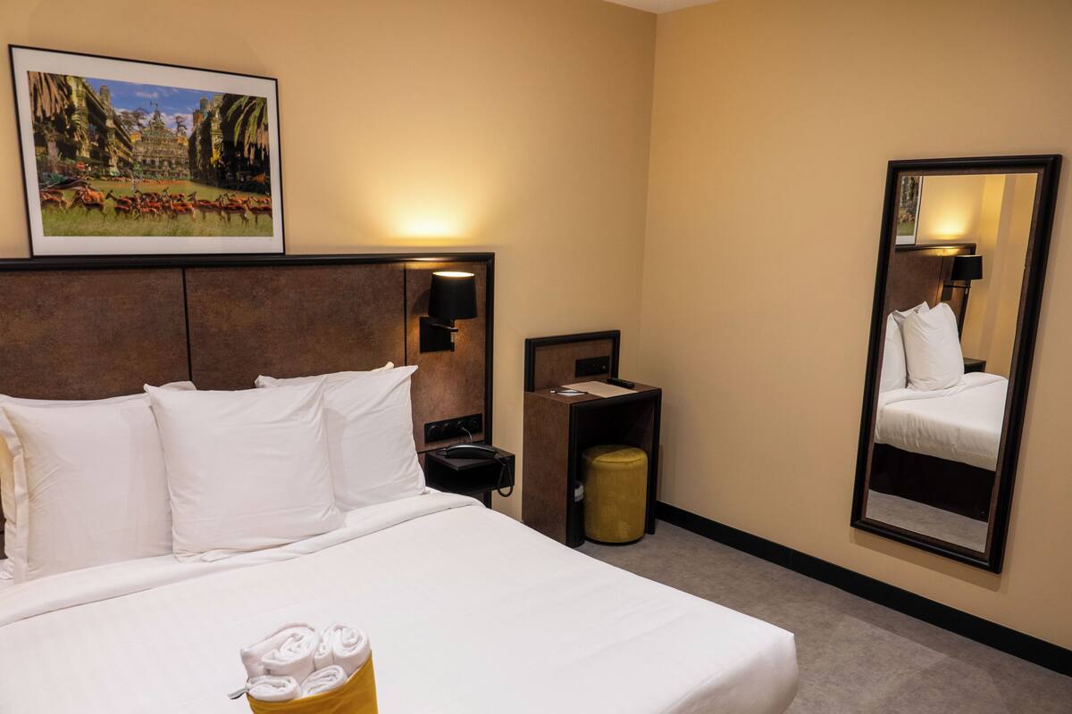A hotel room with a large bed, a bedside table, a mirror, and a wall-mounted picture.