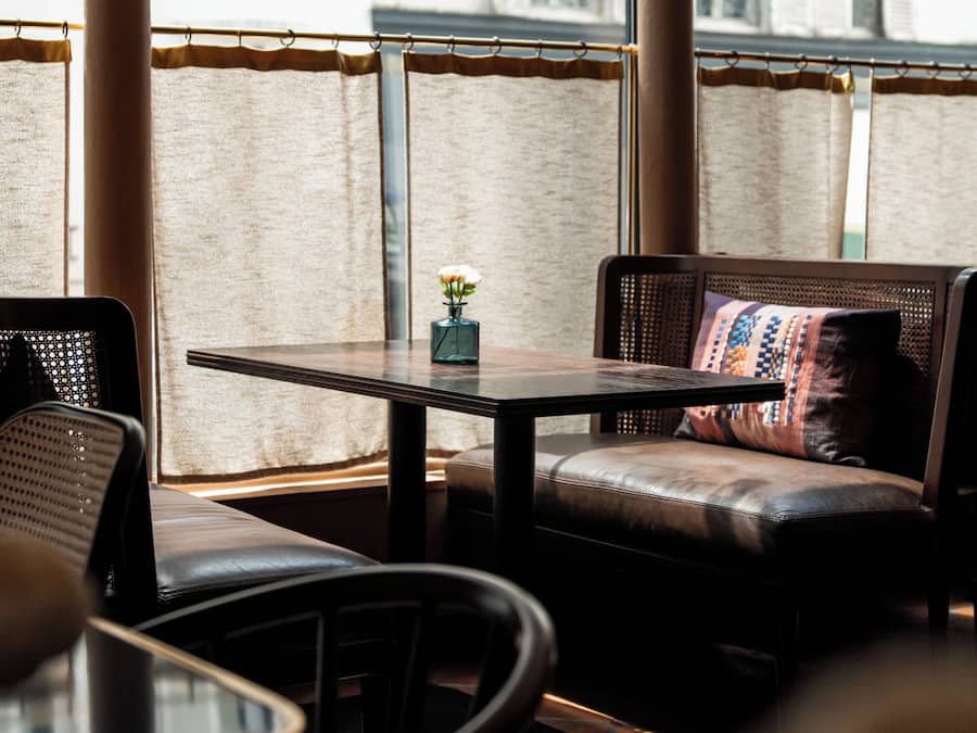 Uma área de estar com mesa e cadeiras, um lustre pendurado, e vista para edifícios através de grandes janelas.