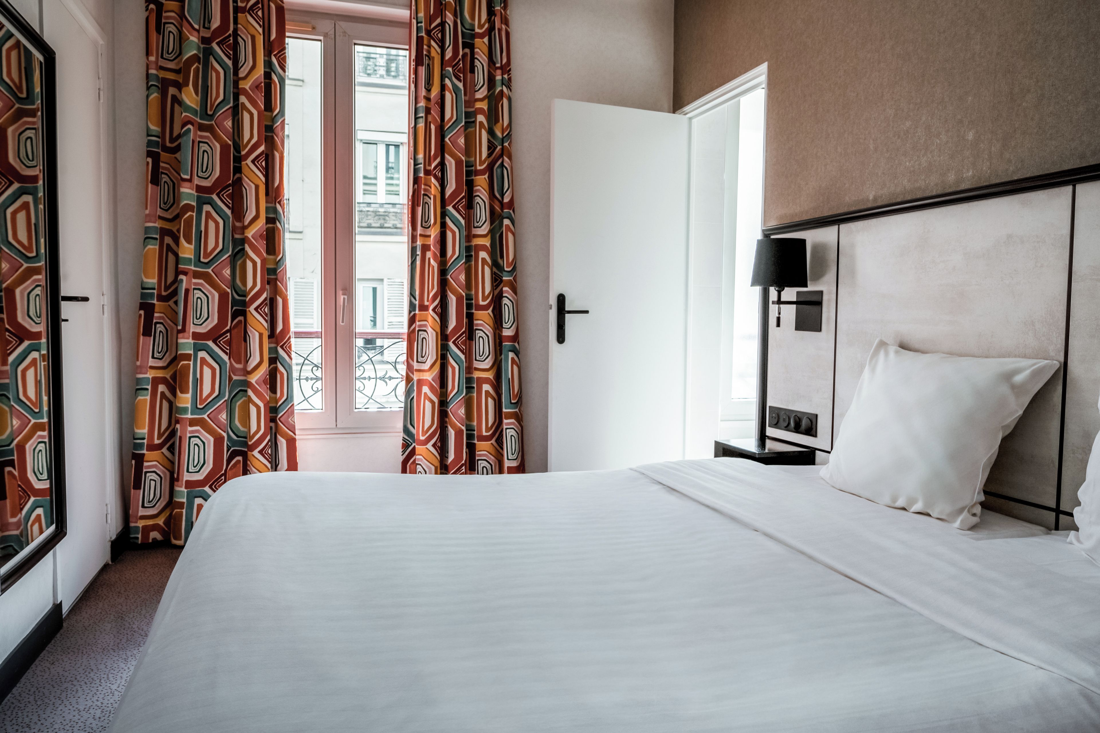 A hotel room with a bed, a nightstand, a lamp, and a view of the outside.