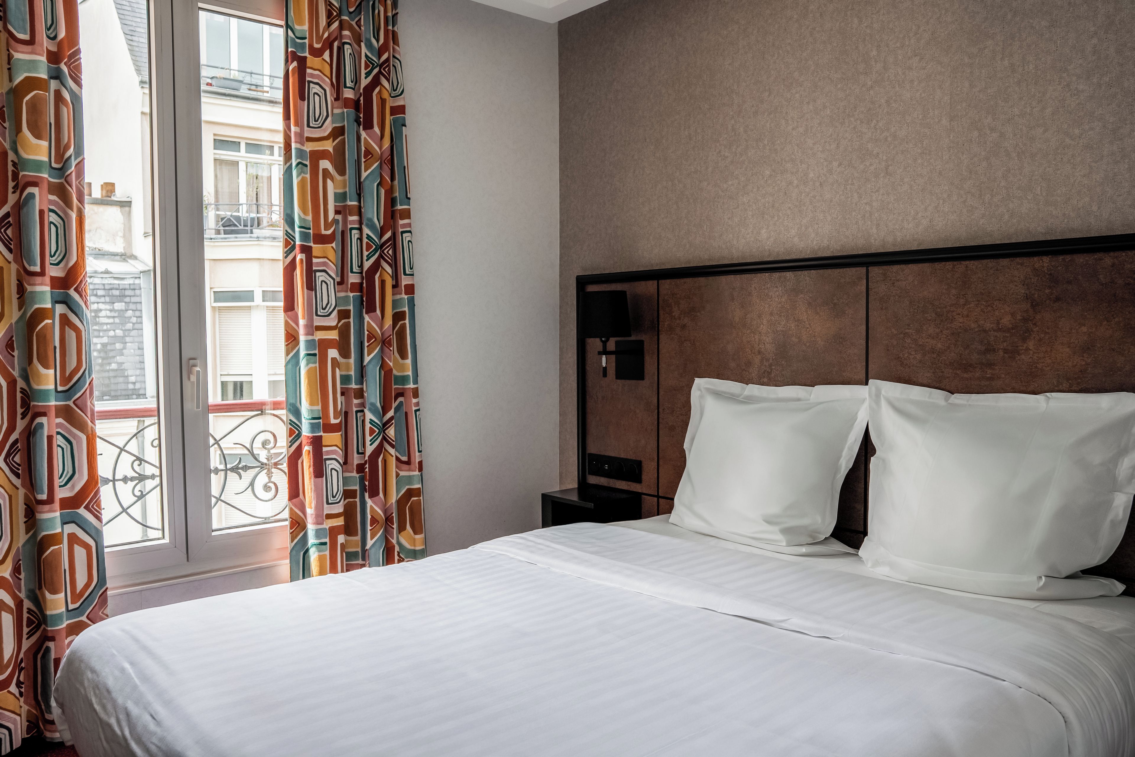 A hotel room with a bed, a nightstand, a lamp, and a view of the outside.