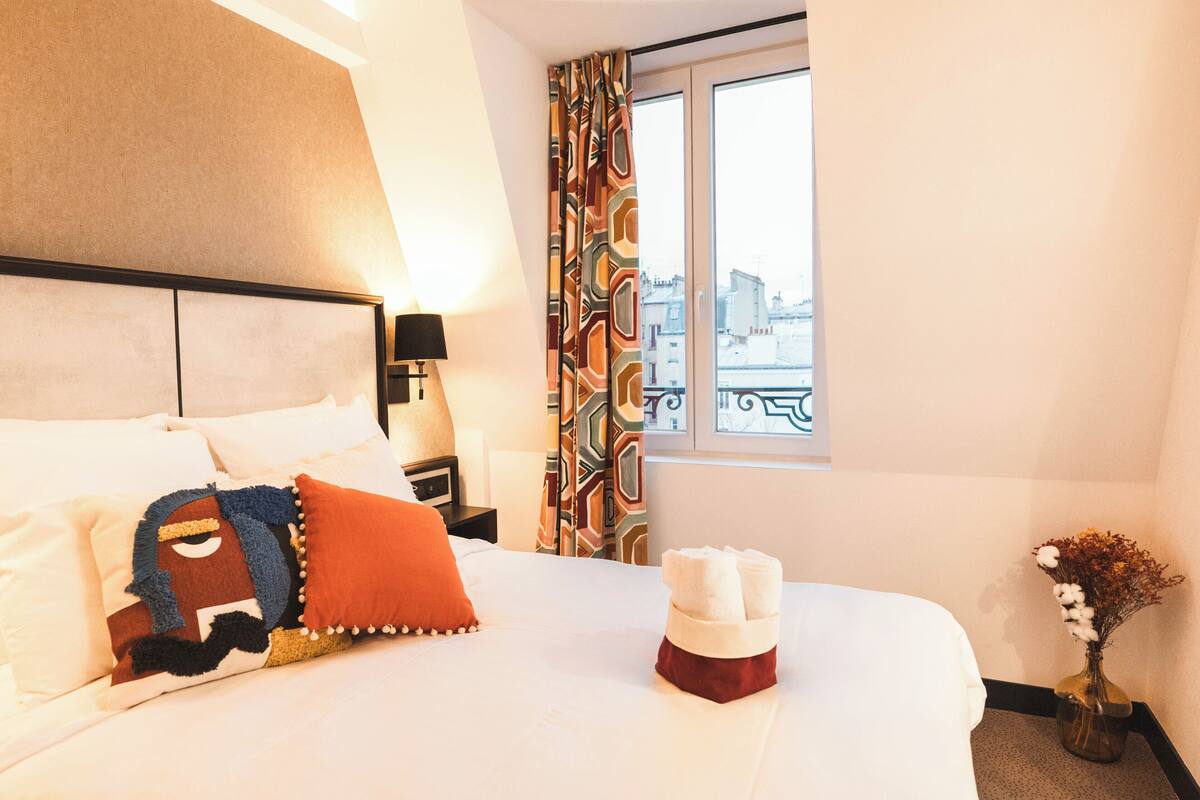 A hotel room with a bed, pillows, a vase with flowers, and a window with a view.